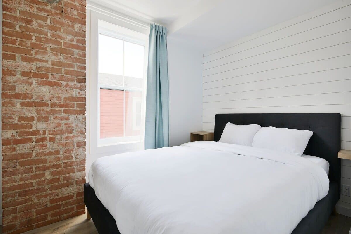 A bedroom with a brick wall, a large window with a curtain, a bed with white bedding, and a wooden nightstand.