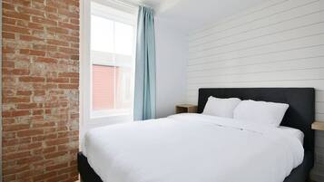 A bedroom with a brick wall, a large window with a curtain, a bed with white bedding, and a wooden nightstand.
