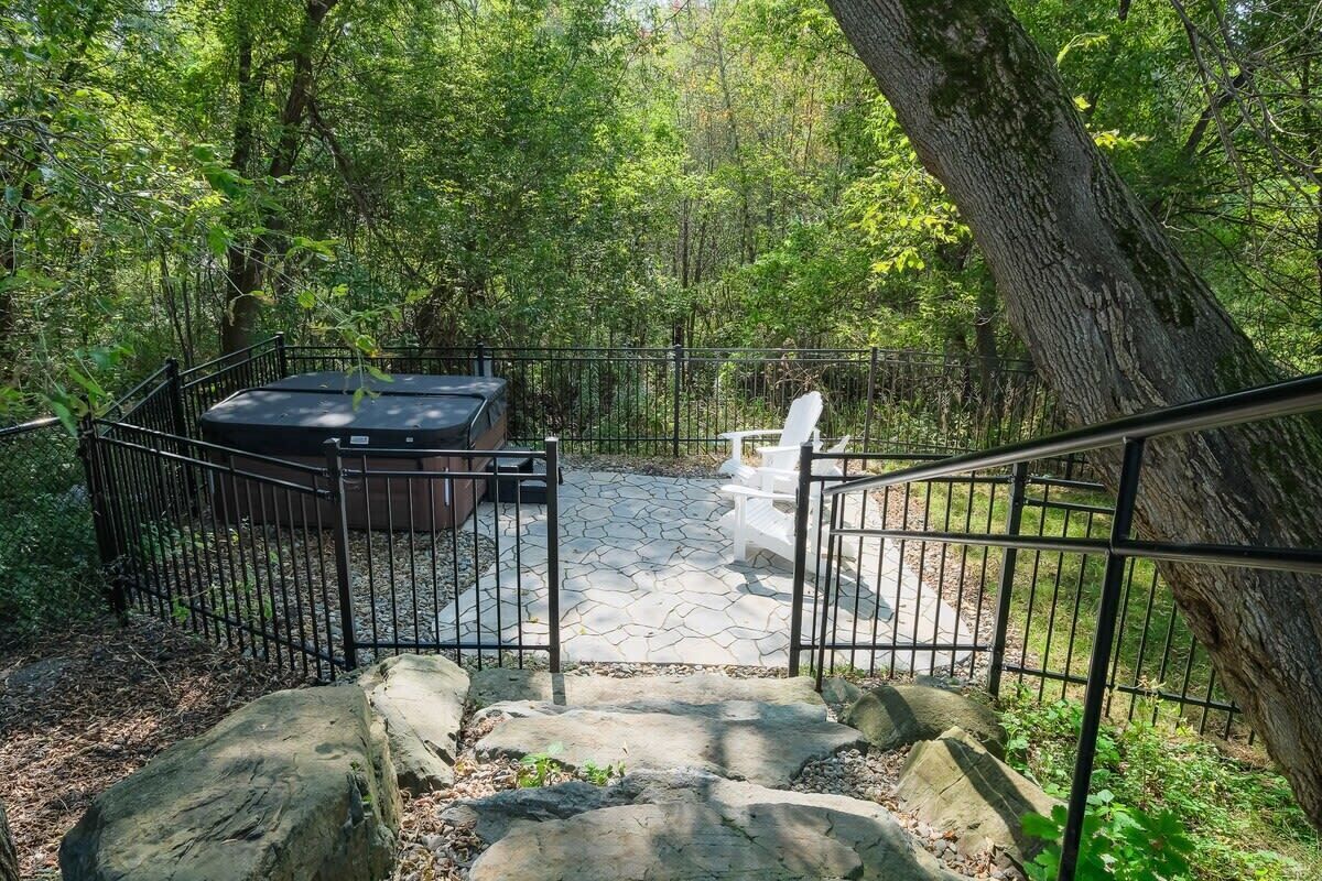 A stone patio with a hot tub, surrounded by a metal fence, and a staircase leading up to a white bench.