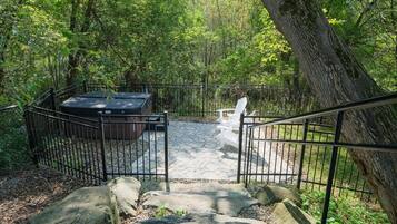 A stone patio with a hot tub, surrounded by a metal fence, and a staircase leading up to a white bench.
