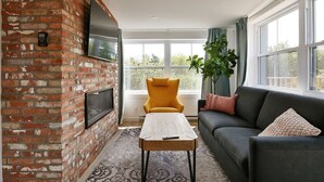 A living room with a brick wall, a flat-screen TV, a yellow armchair, a dark sofa, and a patterned rug. - condo 3 chambres avec foyer,837 shefford suite 200 (Bromont)