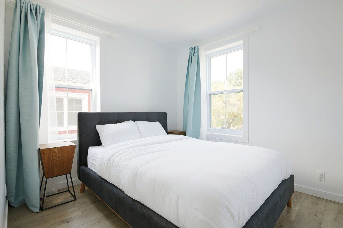 A bedroom with a bed, two windows, a nightstand, and a view of a building outside.