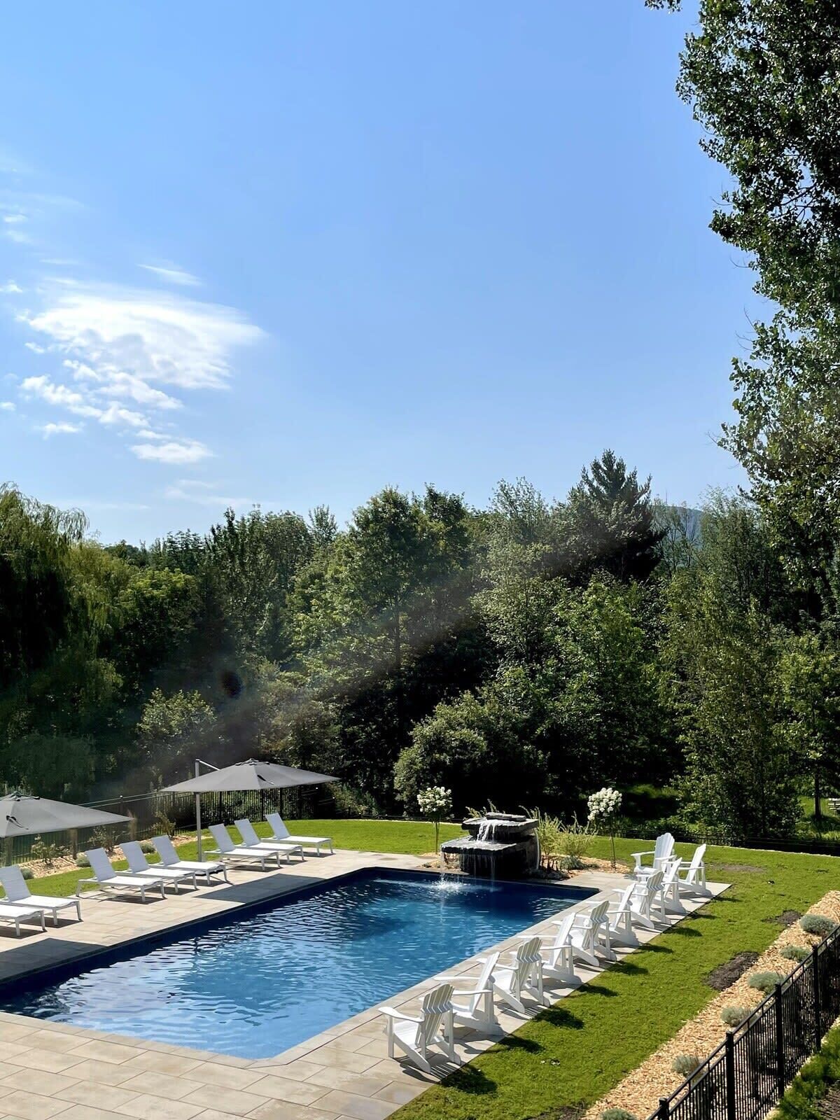 A rectangular swimming pool with a waterfall feature, surrounded by white lounge chairs and a grassy area.