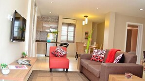Una sala de estar moderna con un sofá marrón, un sillón rojo y una mesa de centro de madera. Se ve una cocina al fondo, con una ventana y una planta.