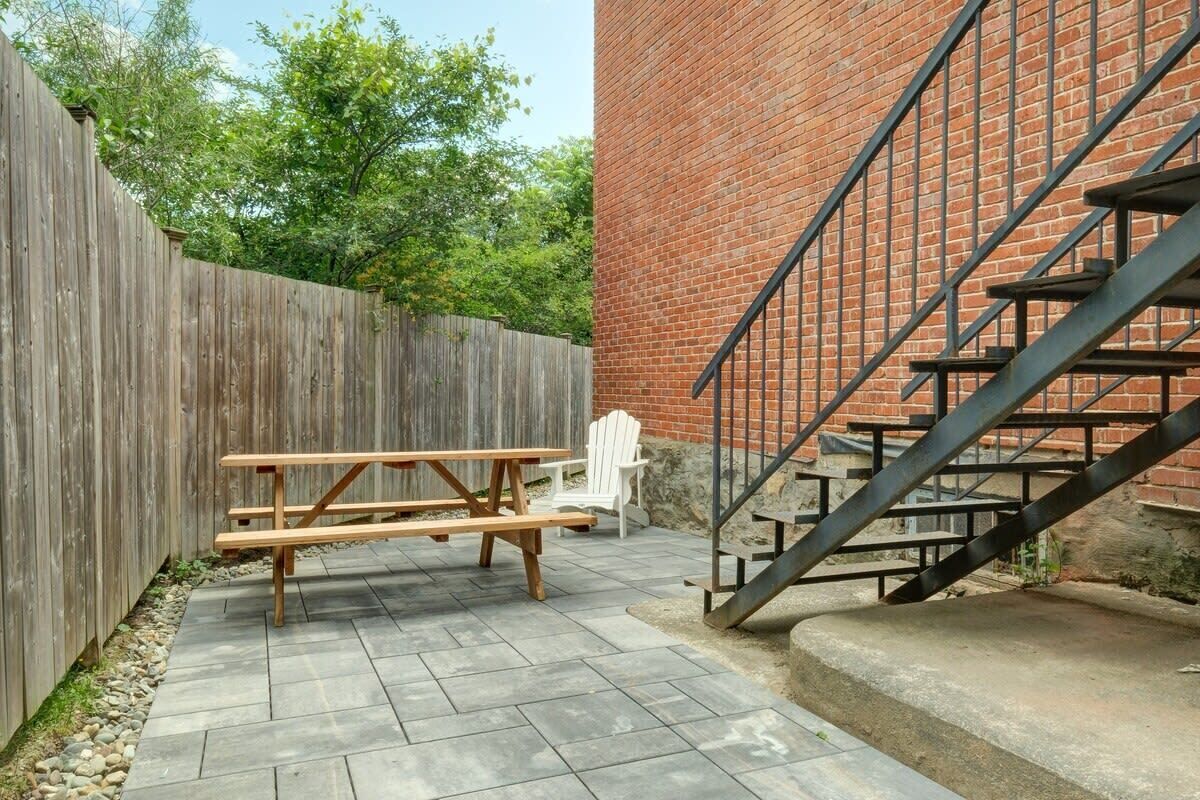 En uteplats med ett träbord för picknick och en vit stol, en mur av tegelsten och en metalltrappa.