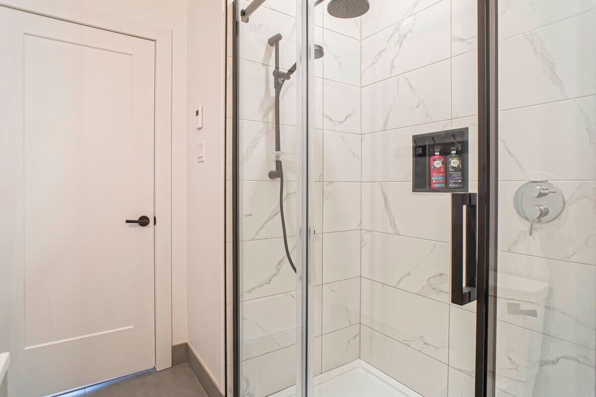 A modern bathroom with a glass-enclosed shower, white marble tiles, and a visible shower control panel.