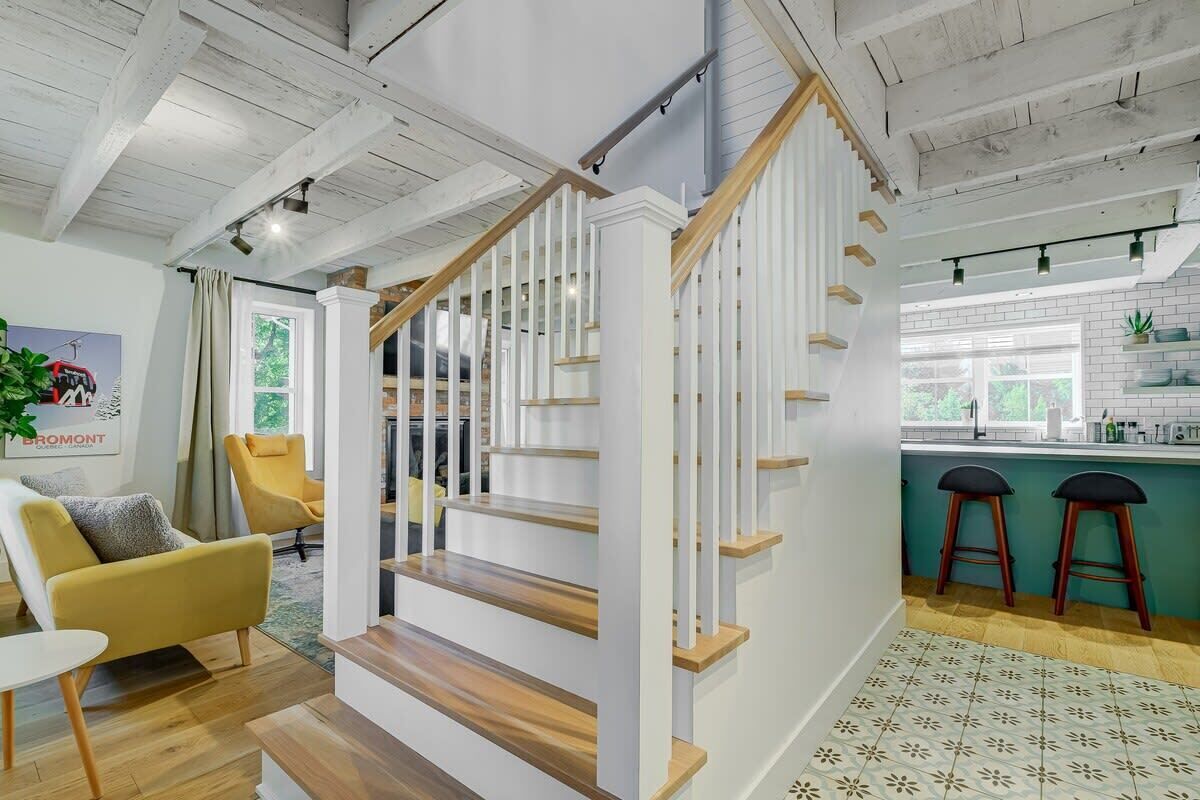 Een modern interieur met een houten trap, een keukenhoek met barstoelen, en een zithoek met een gele fauteuil.