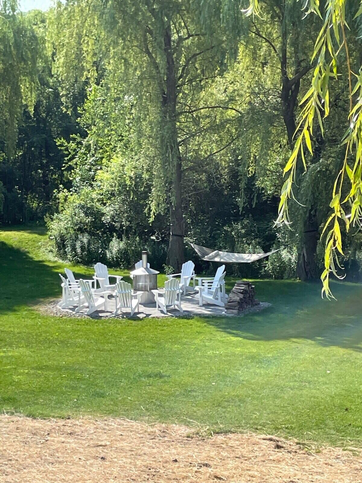 A patio with Adirondack chairs and a wood-burning stove surrounded by lush greenery.