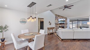 A modern living room with a fireplace, a dining area with a round table and chairs, and a large window overlooking a pool.
