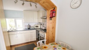 Een keuken met witte kasten, een magnetron, een fornuis en een spoelbak. Er staat een tafel met een zonnebloem tafelkleed en een stoel.