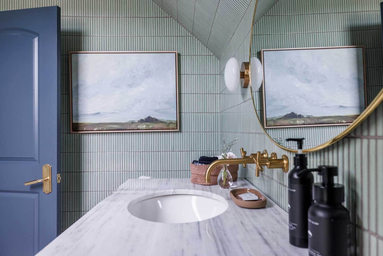 A bathroom with a marble sink, gold fixtures, and two framed landscape paintings on the wall.