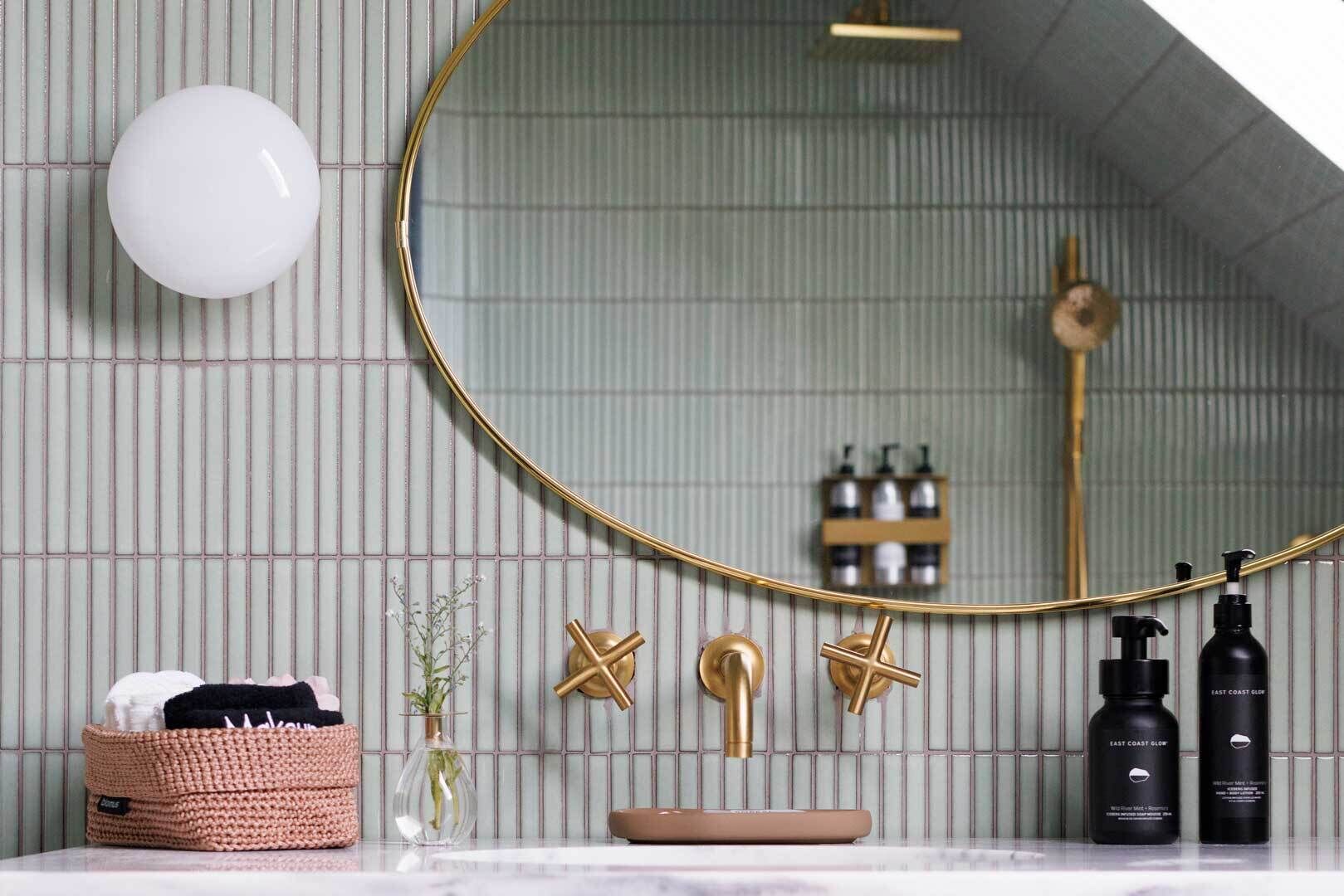 A bathroom with a round mirror, a white light fixture, a starfish-shaped towel holder, a gold faucet, a small vase with flowers, a woven basket with towels, and two black bottles labeled "East Coast Glow".