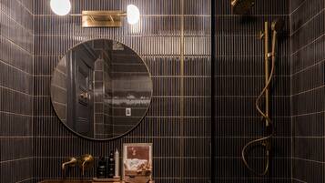 Un baño moderno con una pared de azulejos oscuros, un espejo redondo, un lavabo con grifo dorado y una zona de ducha.