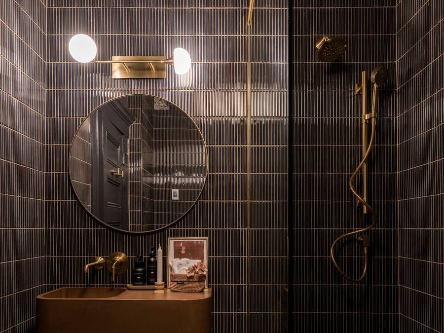 Un baño moderno con una pared de azulejos oscuros, un espejo redondo, un lavabo con grifo dorado y una zona de ducha.
