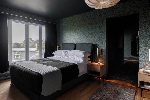 Une chambre moderne avec un grand lit, une table de chevet avec une lampe, une vue sur la ville par la fenêtre et un mur foncé orné d’un motif discret.