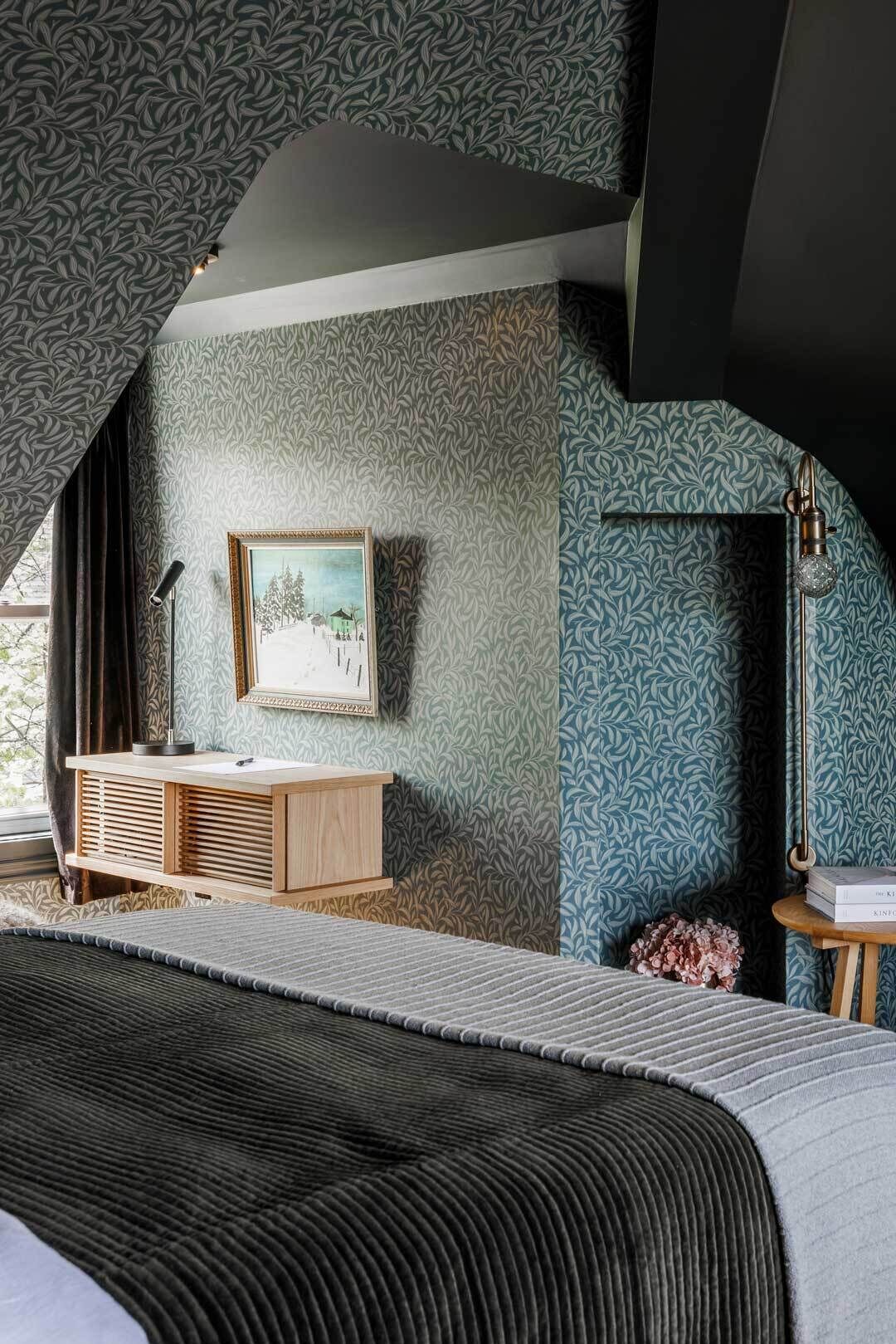 A bedroom with a bed, a wooden dresser, a framed picture, a floor lamp, and a small table with books.
