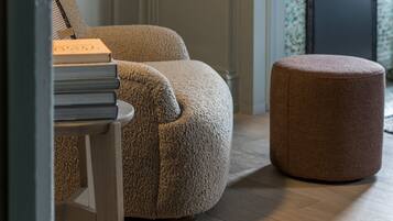 A room with a round, textured ottoman, a small table with books, and a fireplace with a decorative mirror above it.