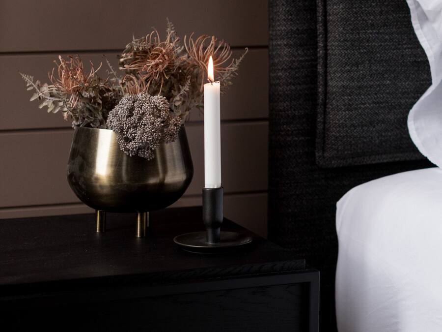 A bedside table with a metallic vase containing dried plants and a lit candle.