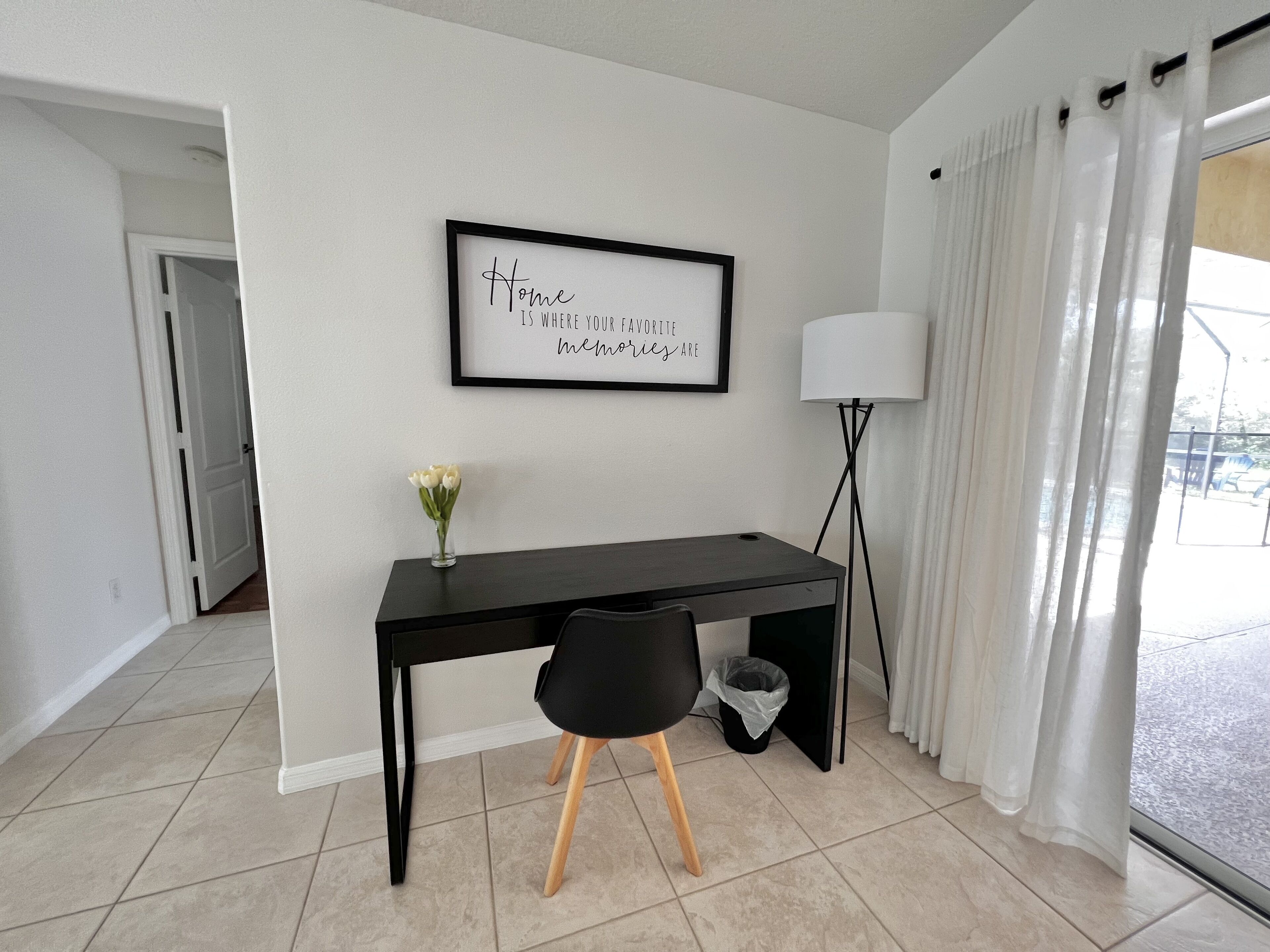 Une pièce moderne avec un bureau noir, une citation encadrée accrochée au mur, un vase rempli de fleurs, une lampe sur pied et une vue sur l’extérieur à travers une fenêtre ornée de rideaux.