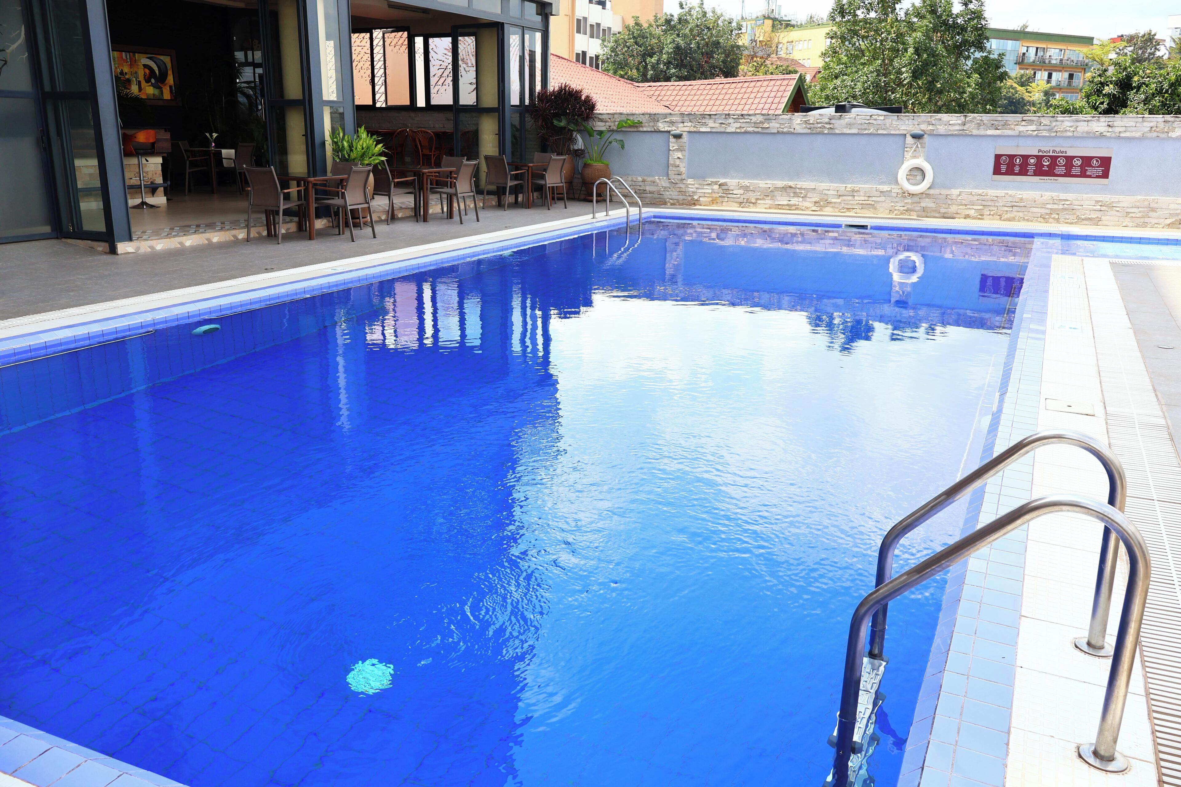 Une piscine aux eaux d’un bleu limpide, avec un escalier en métal sur le côté droit, entourée d’une terrasse carrelée et d’un mur de pierre sur lequel est affiché un panneau indiquant les règles de la piscine.