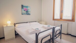 A single bed with a metal frame, white bedding, a bedside table with a lamp, a small bedside drawer, a radiator, and a window with a wooden frame.