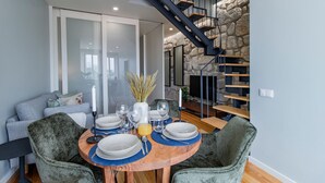 Un coin repas avec une table ronde en bois, dressée pour quatre personnes, entourée de chaises à revêtement vert. Un mur en pierre et un escalier avec des marches en bois sont visibles à l’arrière-plan.