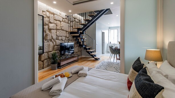 Une chambre moderne avec un mur en pierre, un escalier en bois, un téléviseur à écran plat, un lit avec du linge de lit blanc et, au fond, un coin repas.