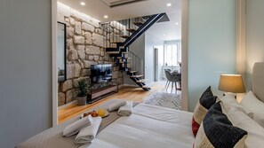 Une chambre moderne avec un mur en pierre, un escalier en bois, un téléviseur à écran plat, un lit avec du linge de lit blanc et, au fond, un coin repas.