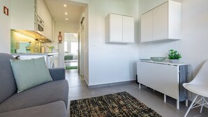 Un espacio habitable moderno y compacto con kitchenette, sofá, comedor y una pequeña mesa con una planta.