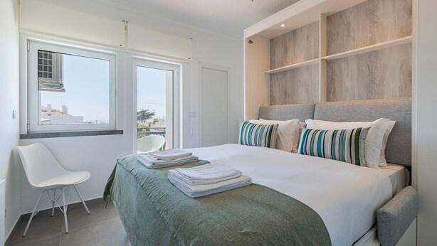 Un dormitorio moderno con una cama grande, una silla y vistas a la ciudad a través de las ventanas.