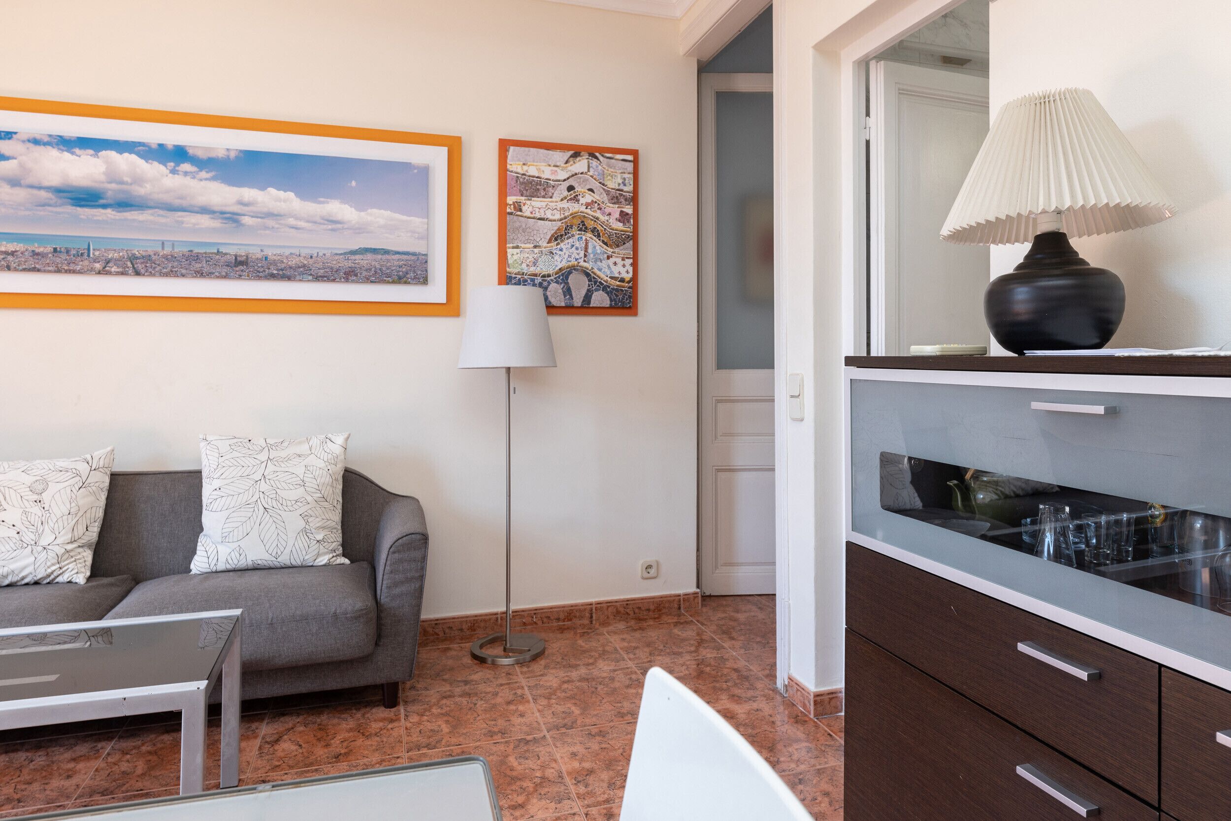 Un salon moderne avec un canapé gris, un lampadaire et un coin repas comprenant une table avec un plateau en verre et des meubles de rangement.