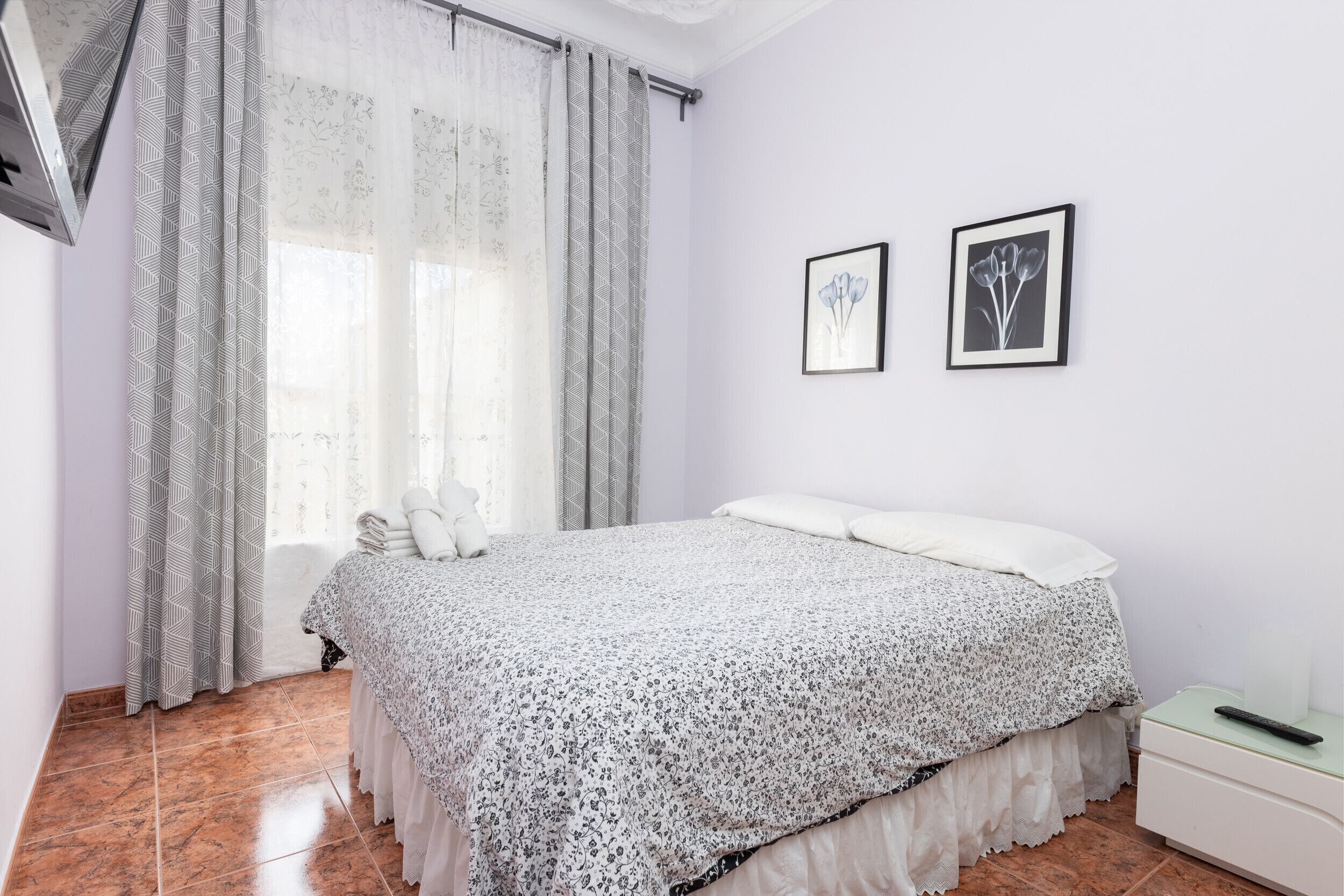 Un dormitorio con cama, mesita de noche, ventana con cortinas y dos cuadros en la pared.