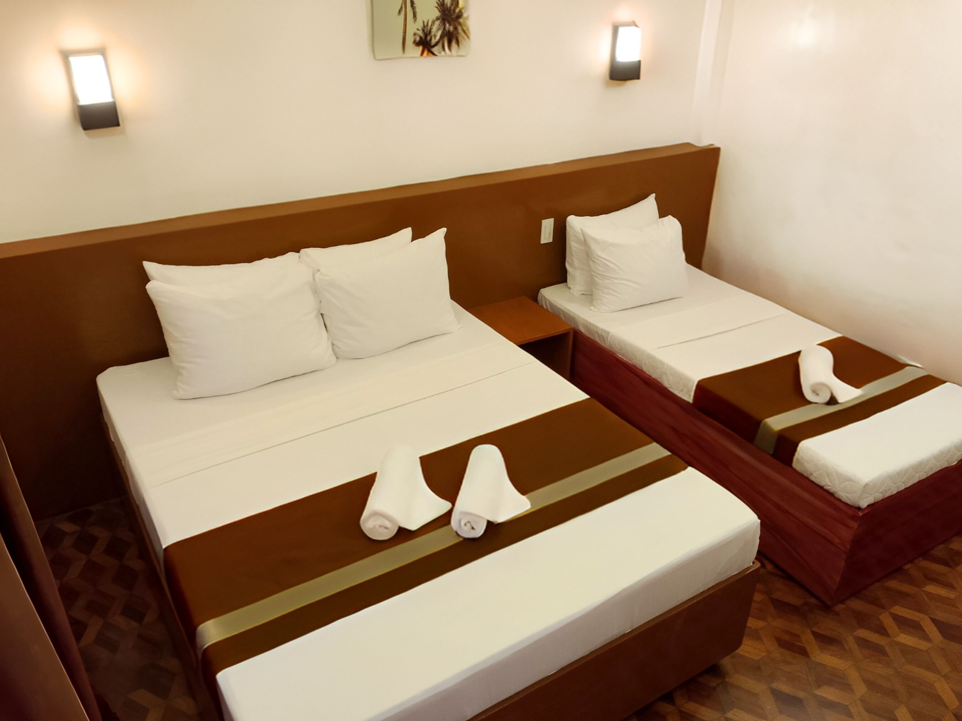 Una habitación de hotel con dos camas individuales, cada una con ropa blanca y cabeceros marrones. Hay dos lámparas de pared y una pequeña decoración en la pared.