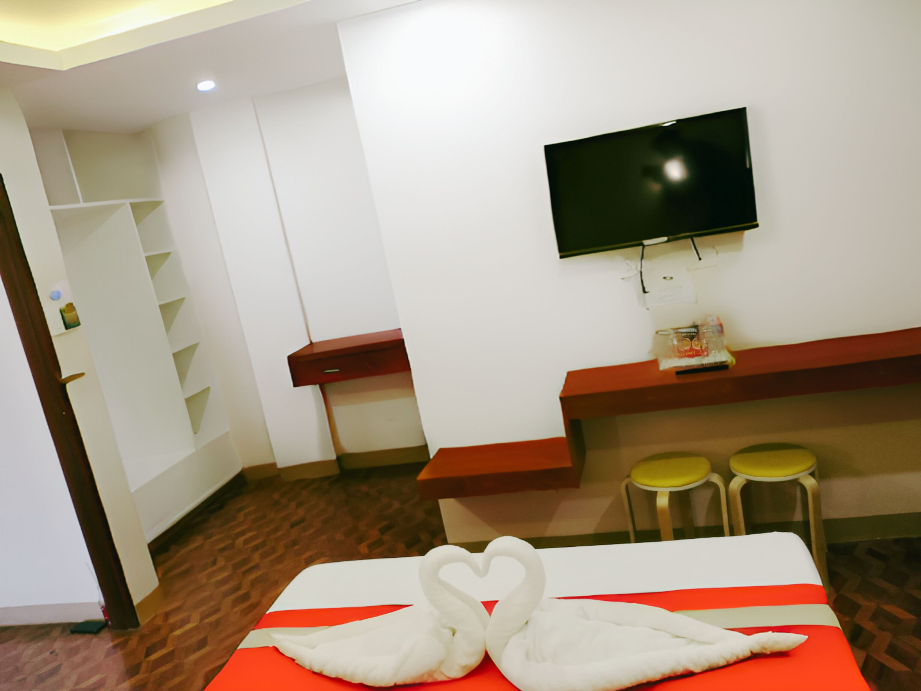 A hotel room with a bed, a TV mounted on the wall, a desk, and two stools.