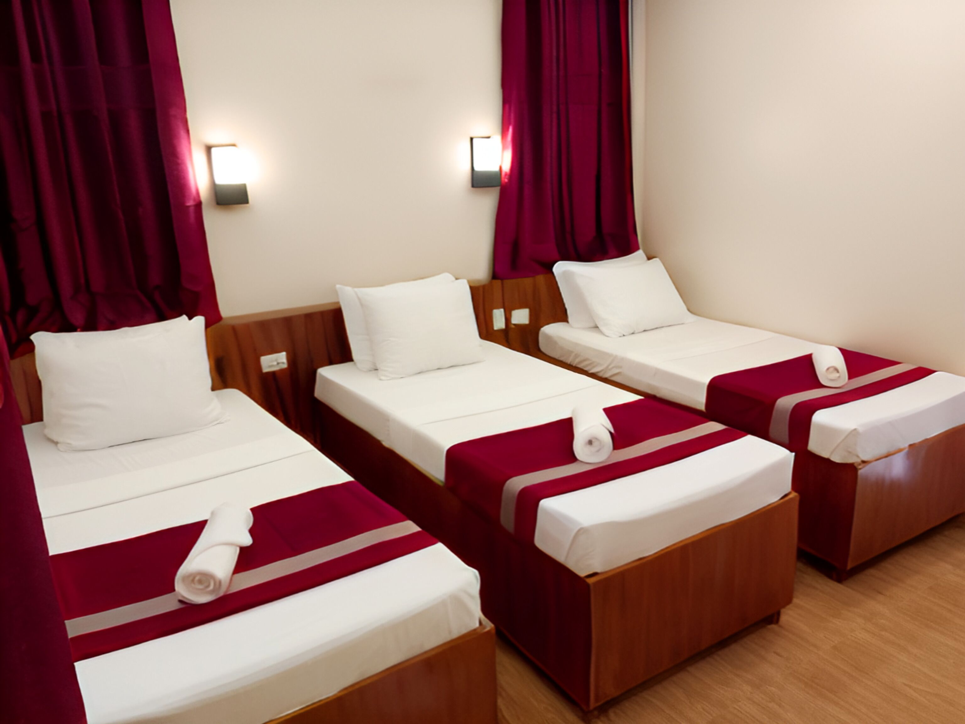 A hotel room with three single beds, each with a wooden headboard, white bedding, and a red accent.