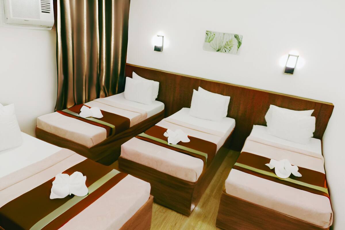 Habitación de hotel con cuatro camas individuales, cada una con cabecero de madera y almohada blanca. Se observa un equipo de aire acondicionado instalado en la pared y un pequeño cuadro enmarcado.