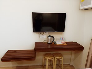 A compact room with a flat-screen TV mounted on the wall, a wooden desk with a kettle, two stools, and a small table with a tray of items. - Dy Heritage Suites (Vigan)
