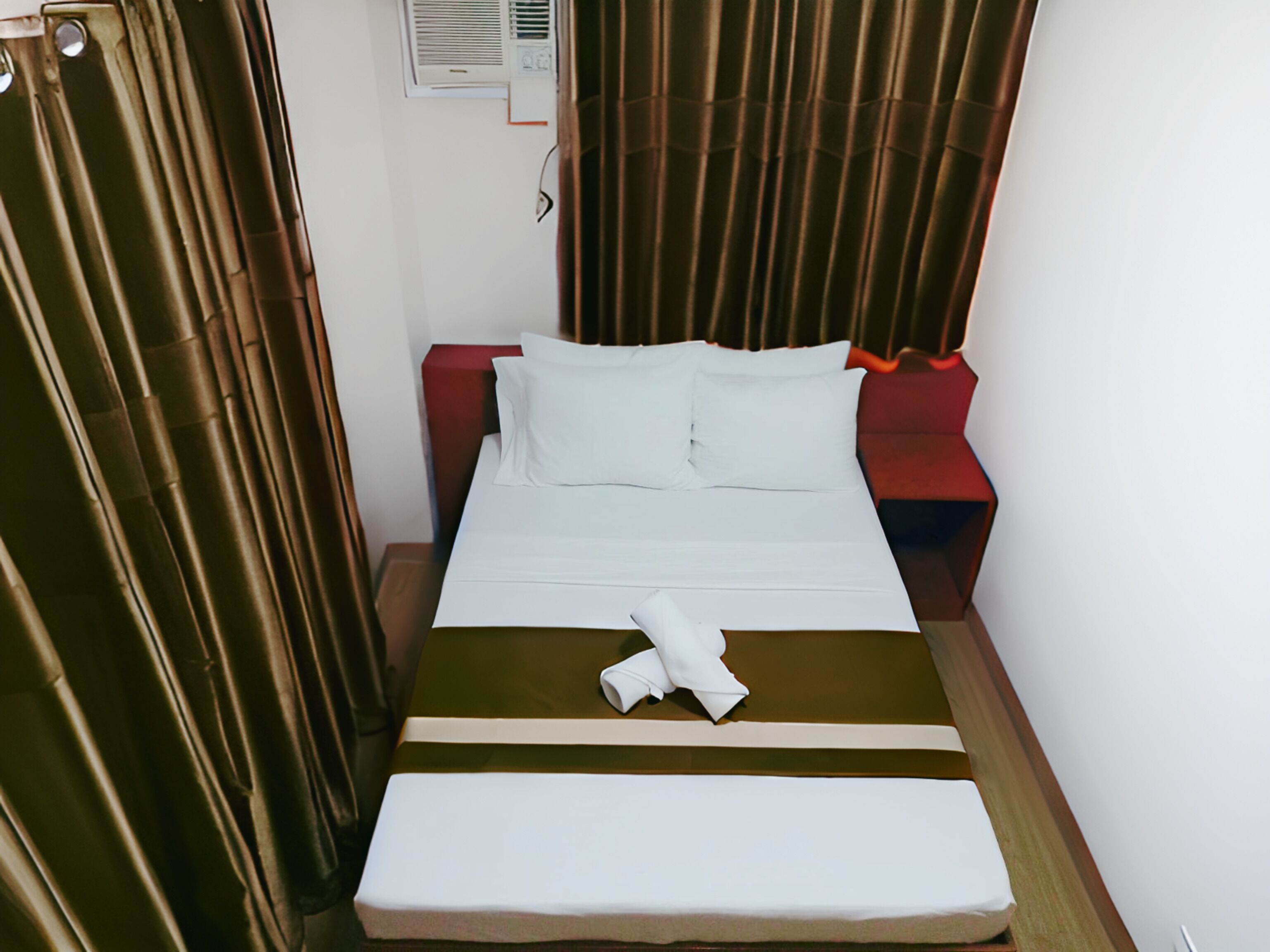 A hotel room with a bed, a red nightstand, a white pillow, and a brown curtain.