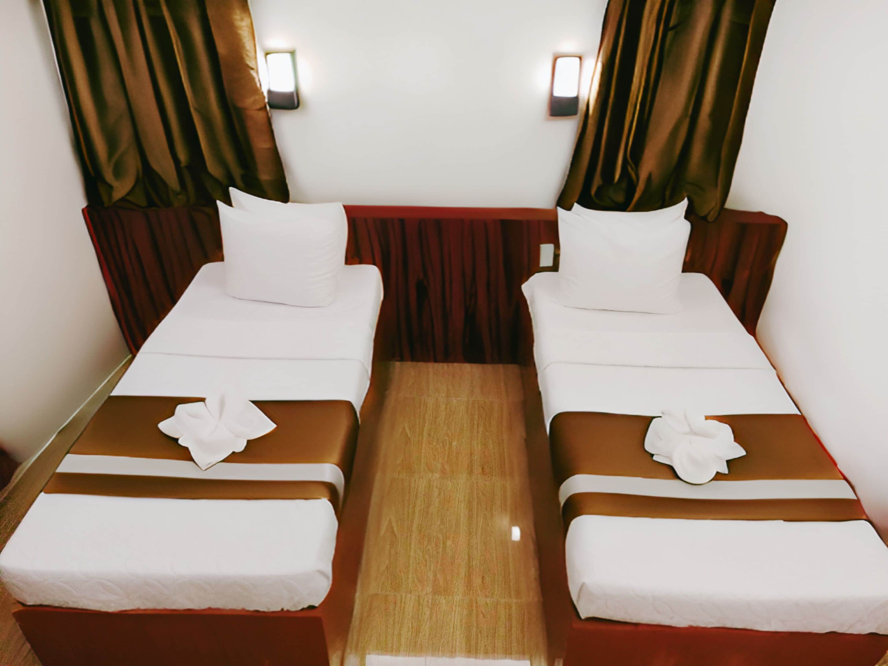 A hotel room with two single beds, each with a white pillow and a decorative white flower. The beds have brown headboards and footboards, and there is a wooden table between them.