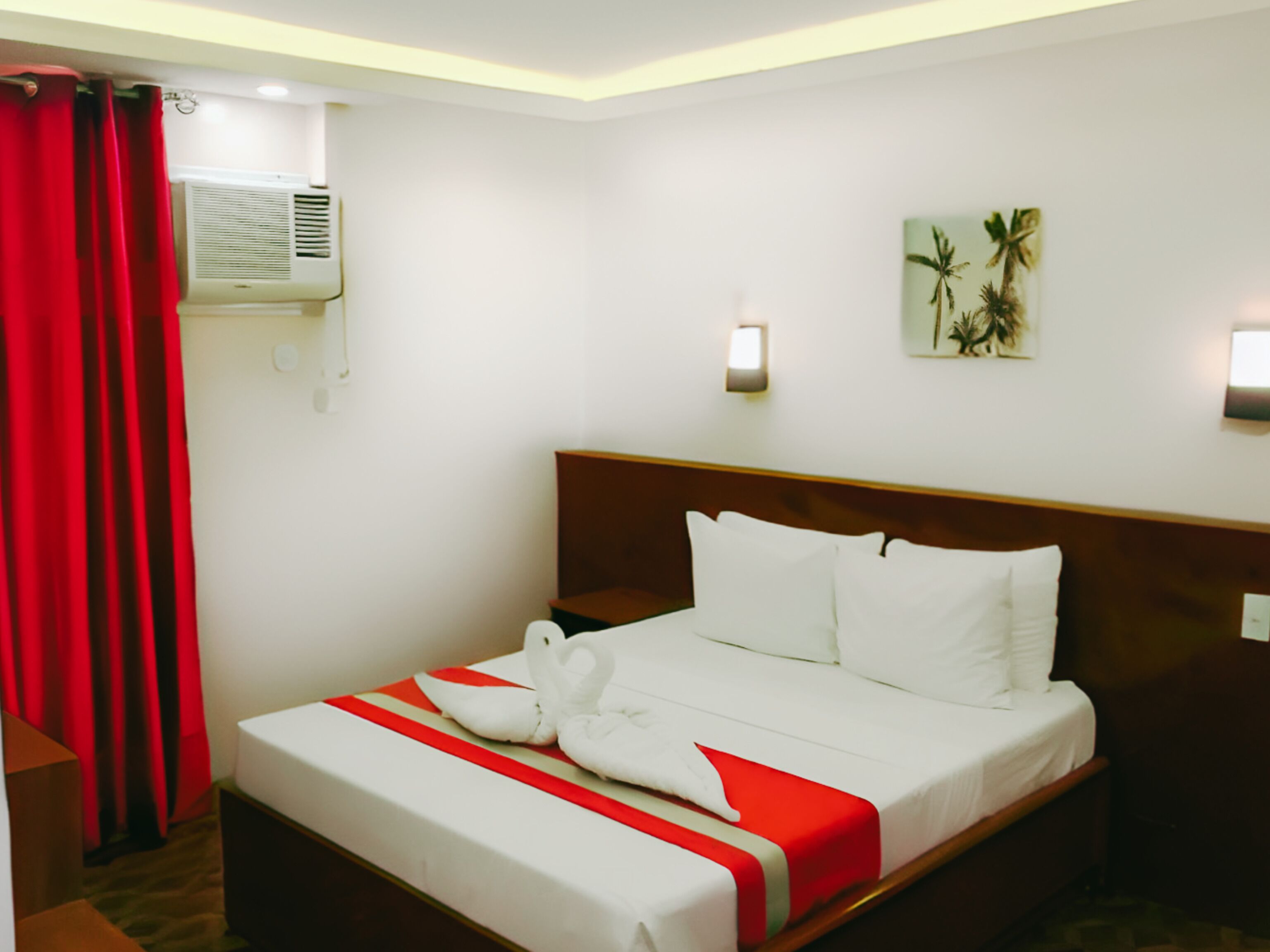 A hotel room with a bed, red curtains, an air conditioning unit, a wall-mounted light, and a palm tree painting.