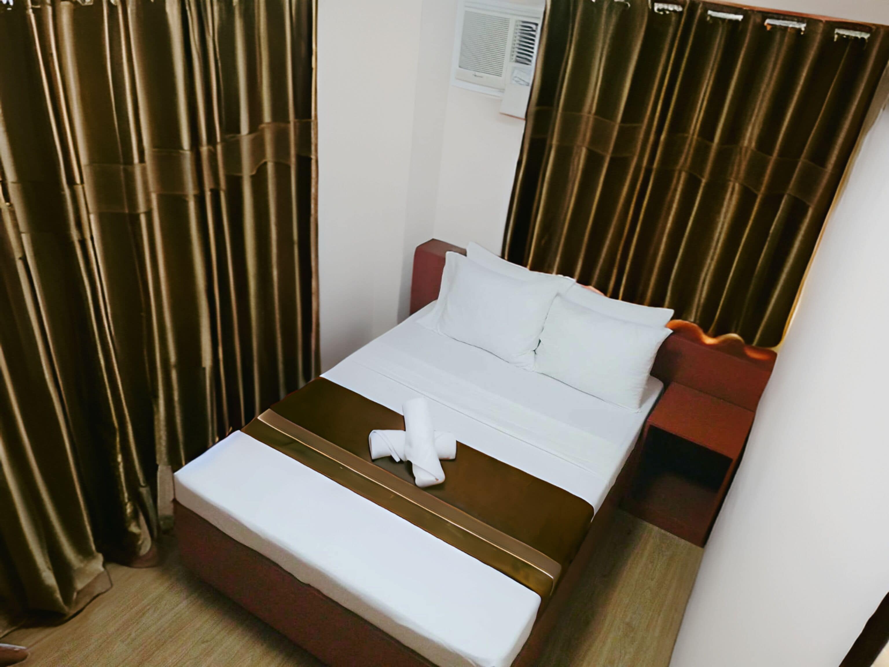 A neatly made bed with white linens and a brown headboard, a bedside table with a red accent, and a window with blinds.