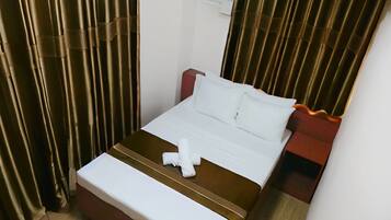 A neatly made bed with white linens and a brown headboard, a bedside table with a red accent, and a window with blinds.