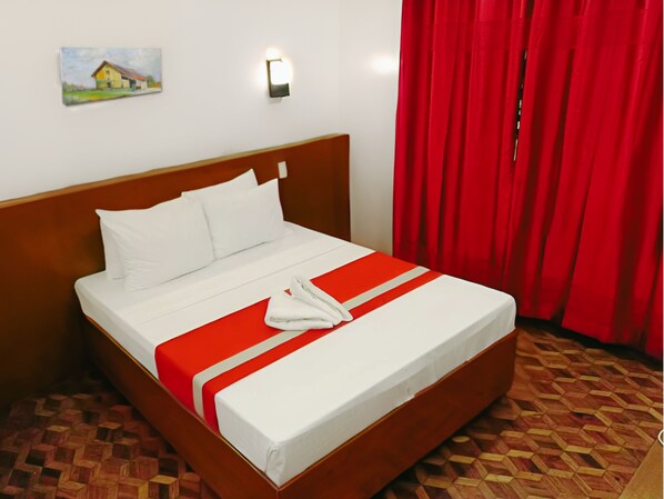 Una cama bien tendida con ropa blanca y un detalle rojo, un cabecero de madera, un cuadro en la pared y cortinas rojas.