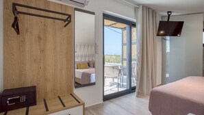 A modern bedroom with a wooden headboard, a bedside table with a safe, a large mirror, a flat-screen TV, and a balcony with a view.