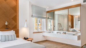 A modern hotel room with a large bed, a vanity with a mirror, a small table with a lamp, and a window with curtains.