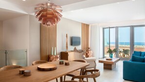 A modern living room with a wooden dining table, chairs, and a blue sofa. There is a large window with a view of the beach.