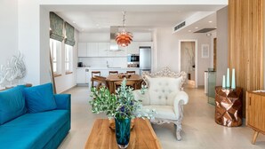 A modern living room with a blue sofa, a wooden coffee table, and a white armchair. There is a kitchen in the background with white cabinets and a dining area with a wooden table and chairs.