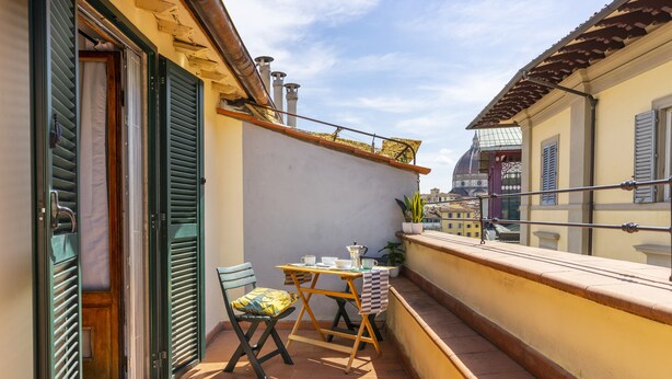 Een klein balkon buiten met een tafel en stoelen, een bloempot en uitzicht op de stad.