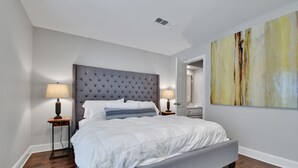 Une chambre avec un grand lit, deux tables de chevet avec des lampes, une tête de lit capitonnée, un grand tableau abstrait et un plancher en bois.