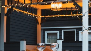 Una mesa con dos copas de vino vacías y una silla, frente a la fachada de una casa con luces colgantes y una ventana.